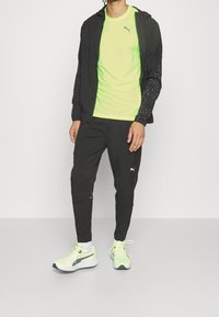 Chaqueta atlética negra con mangas texturizadas, camiseta verde neón, pantalones negros y zapatillas verde claro con detalles en negro.