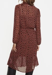 Brązowa przezroczysta sukienka w czarne kropki, z długimi rękawami, falbaną przy kołnierzyku, zwężoną talią i swobodnym dołem.