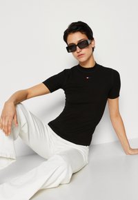 Haut noir à manches courtes avec un col haut et un discret logo rouge. Le mannequin porte un pantalon large blanc et des lunettes de soleil noires surdimensionnées.