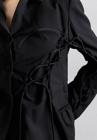 Schwarze Jacke mit strukturiertem Design, einem Reißverschluss-Brusttasche und aufwendigen Schnürdetails an den Ärmeln für zusätzliche Textur.
