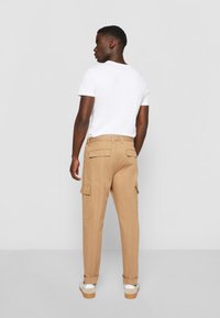 Beige Cargo-Hosen mit Rücken- und Seitentaschen, schmal geschnitten, aus Baumwollmaterial, kombiniert mit einem weißen T-Shirt und weißen Sneakern.