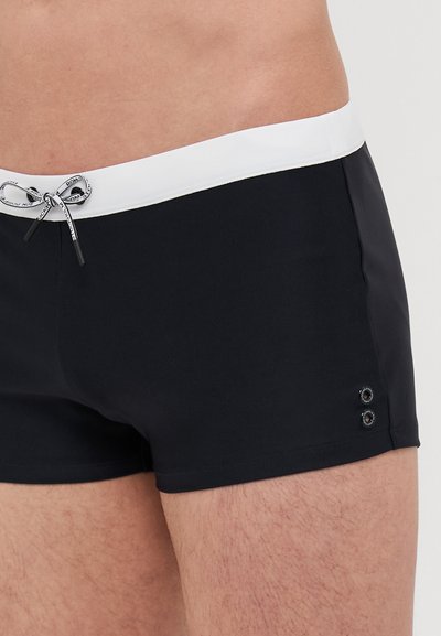 Shorts de bain noirs avec une ceinture élastique blanche, fermeture à cordon, et deux œillets argentés sur le côté. Matériau lisse et extensible.