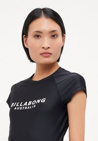 Μαύρο αθλητικό μπλουζάκι με κοντά μανίκια, φτιαγμένο από λείο ύφασμα, που διαθέτει λευκό λογότυπο "BILLABONG AUSTRALIA" στο στήθος.