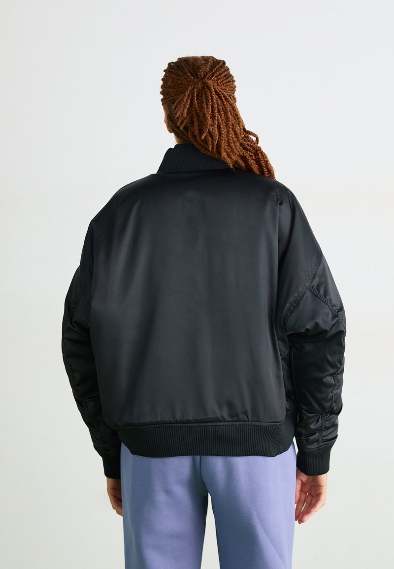Veste bomber noire avec un col et des poignets côtelés, présentant une texture lisse et brillante ainsi que des épaules rembourrées, portée sur un pantalon lilas.