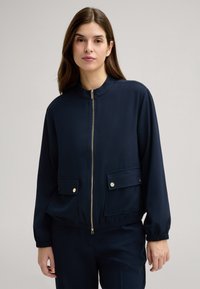 Marineblaue Jacke mit hohem Kragen, Reißverschluss, zwei vordere Taschen mit Knopfakzenten und elastischen Bündchen. Glatte Stofftextur.