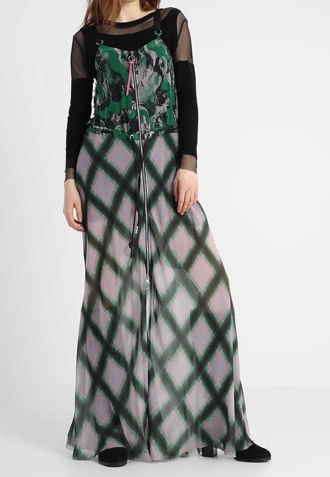 Femme portant une longue robe verte et rose à motifs avec des détails de fermeture éclair, par-dessus un haut noir transparent à manches longues, se tenant contre un fond blanc.