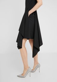 Femme en robe jupe asymétrique noire, main dans la poche, portant des chaussures à talons hauts pointues argentées et scintillantes, debout sur un sol gris.