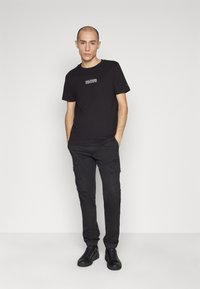 T-shirt nera in cotone con un logo sul petto, abbinata a pantaloni cargo neri con tasche laterali e sneakers nere.
