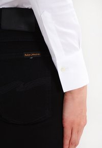 Camisa blanca de manga larga con puño puntiagudo y detalle de botón, combinada con jeans de mezclilla negros que presentan una etiqueta y costuras sutiles en el bolsillo.