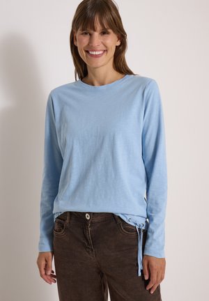 Femme souriante portant une chemise bleu clair à manches longues avec des liens latéraux et un pantalon marron foncé sur un fond clair uni.