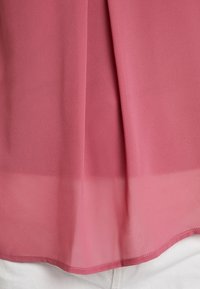 Ljusrosa blus i genomskinligt tyg med en något texturerad yta, med en rynkad design vid halsringningen och en rundad nederkant.