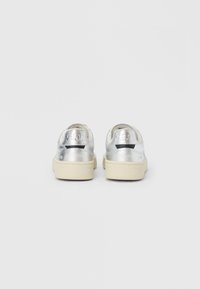 Hopeanväriset metallinhohtoiset tennarit, joissa on sileä pinta, kerma valkoinen kumipohja ja kohokuvioitu logo kannassa. Minimalistinen muotoilu pehmeillä yksityiskohdilla.