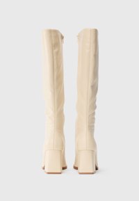 Bottes en cuir crème jusqu'aux genoux avec des talons carrés, des fermetures éclair arrière visibles et de subtils détails de couture sur un fond blanc uni.