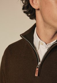 Brauner Sweater mit Reißverschluss und strukturiertem Finish, versehen mit einem silbernen Reißverschluss und einem Leder-Ziehband, übereinander getragen mit einem weißen Hemd mit Knöpfen.