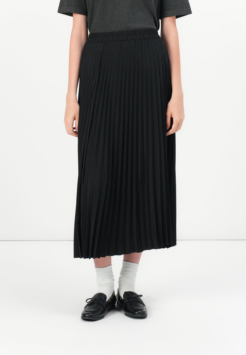 Gonna midi a pieghe nera con vita elastica, caratterizzata da sottili pieghe verticali e una texture liscia, abbinata a scarpe nere e calzini bianchi.