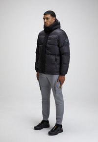 Giacca a piumino nera con cappuccio, dotata di chiusura con zip e tasche laterali. Pantaloni cargo grigi con tasche laterali e marchio. Scarpe sportive nere.