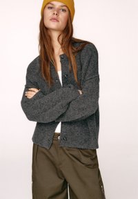 Šedý pletený kardigán s okrúhlym výstrihom, rebrovanými manžetami a zapínaním na gombíky vpredu, skombinovaný s olivovo zelenými nohavicami. Minimalistický dizajn.