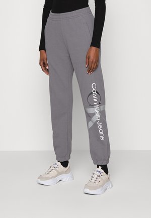 Henkilö, jolla on harmaat Calvin Klein Jeansin collegehousut joustavilla resoreilla, musta yläosa ja beigen väriset paksupohjaiset tennarit yksiväristä taustaa vasten.
