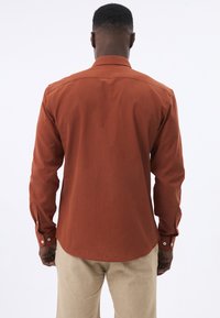 Camisa de manga larga color óxido con puños abotonados, un cuello clásico y un dobladillo curvado. La tela parece suave con una ligera textura.