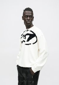 Άνδρας με κοτσίδες φοράει λευκό φούτερ με μεγάλο μαύρο αφηρημένο σχέδιο και μαύρο δερμάτινο παντελόνι, τα χέρια στις τσέπες, κοιτώντας πλάγια.