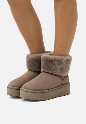 Botas cortas marrones hasta el tobillo, hechas de ante, con un suave puño de piel sintética gris, que cuentan con una suela plana de goma y un diseño de punta redondeada.