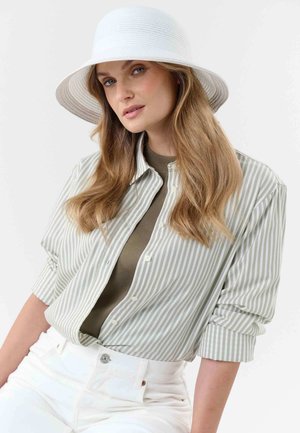Woman with long blonde hair wearing a white wide-brim hat, green striped button-up shirt over olive top, and high-waisted white pants.