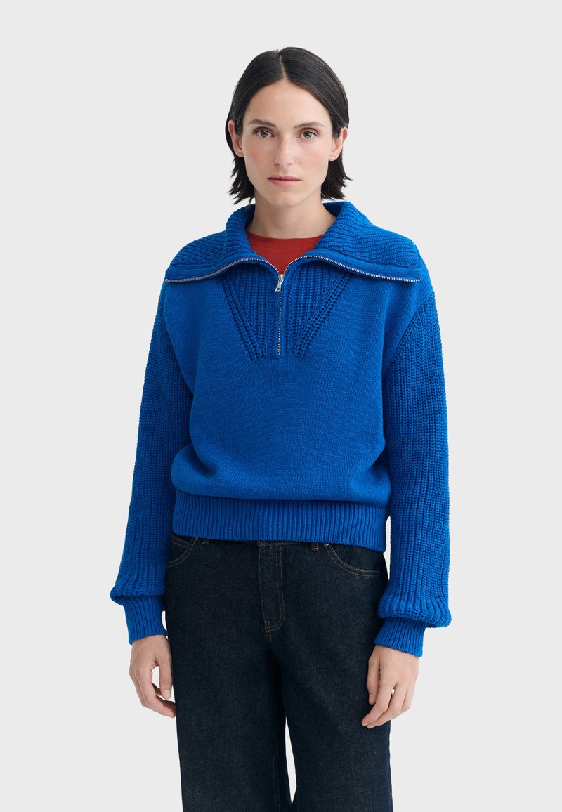 Femme portant un pull en tricot bleu vif avec une fermeture éclair à moitié et une chemise rouge en dessous, associée à un jean bleu foncé.
