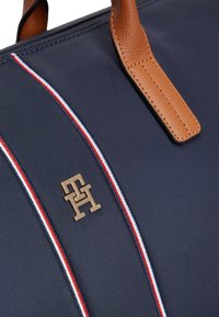 Τσάντα ώμου από ναυτικό νάιλον με καφέ δερμάτινες χειρολαβές, διακοσμημένη με χρυσό λογότυπο και κόκκινες, λευκές και μπλε ριγέ λεπτομέρειες κατά μήκος των ραφών.