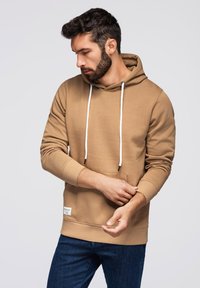 Hnědá mikina z měkkého materiálu, s klokankou, nastavitelnými šňůrkami a ribbovanými manžetami a lemem. Nosená s modrými džíny.