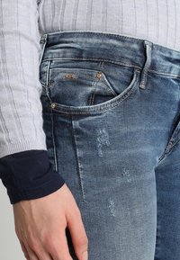 Modré džíny z denimu s opotřebovaným vzhledem, se klasickým pětipocket designem a viditelným opotřebením na přední straně. Dekorativní šití blízko pasu.