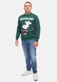 Maglione verde lavorato a maglia con Snoopy nella veste di chef, con la scritta "MASTER CHEF". Indossato con jeans blu e sneakers bianche.