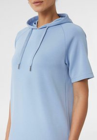 Jasnoniebieska bluza z kapturem z krótkimi rękawami, wykonana z miękkiego materiału, z detalami w postaci sznurka i metalowymi końcówkami w kolorze srebra. Charakteryzuje się luźnym krojem i gładką fakturą.
