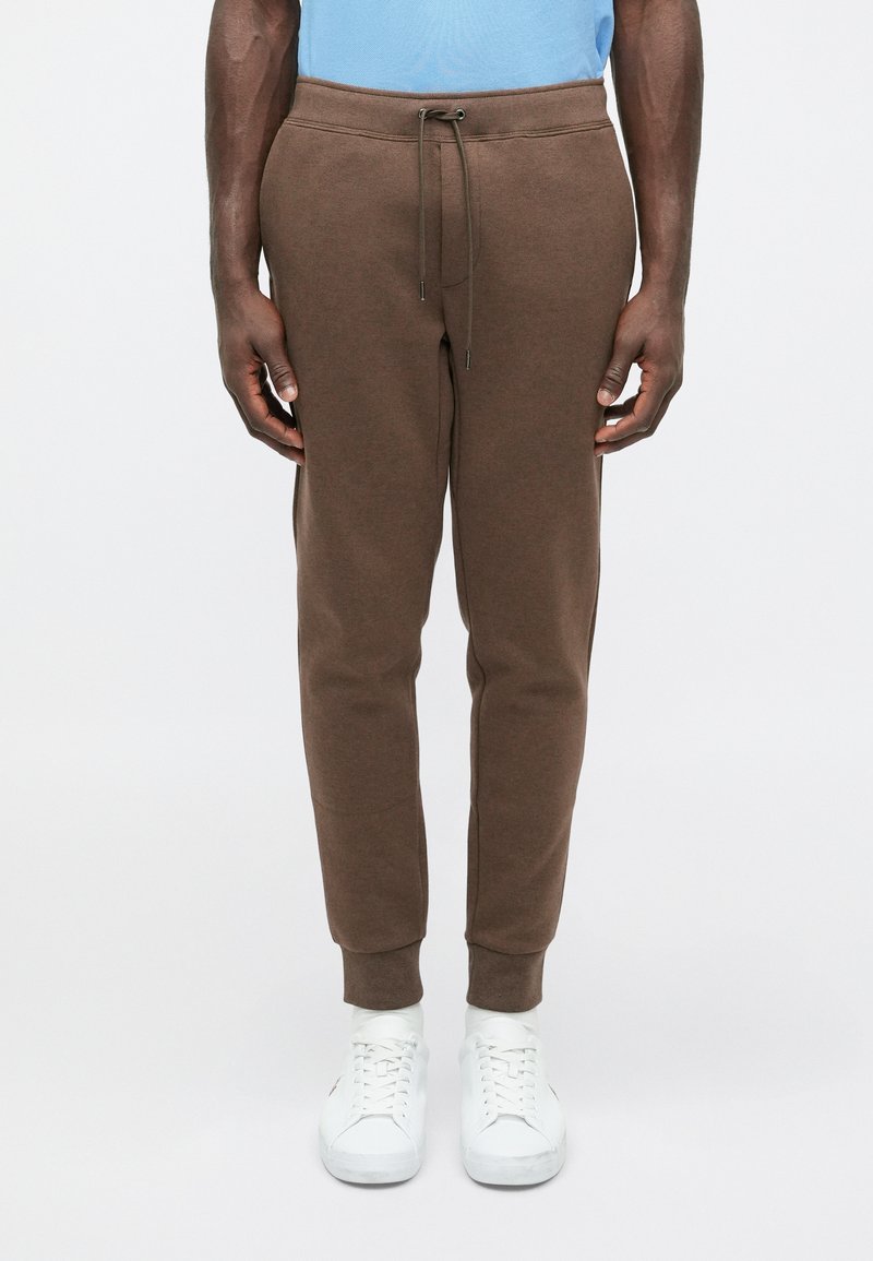 Καφέ βαμβακερές φόρμες με κορδόνι στη μέση, στενές στα πόδια και ραμμένα μανίκια, σε συνδυασμό με λευκά αθλητικά παπούτσια. Απαλή υφή, ελάχιστες λεπτομέρειες.