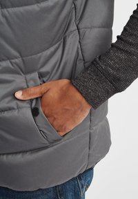 Graue Steppweste mit einer Seitentasche, die eine glatte Textur und subtile Nähte aufweist. Die Hand befindet sich in der Tasche.