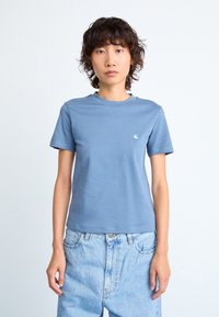 Person iført en lyseblå t-skjorte med korte ermer og lyseblå høy midje jeans som står foran en ensfarget hvit bakgrunn.