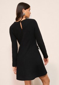Rochie tricotată neagră cu mâneci lungi, având un detaliu în formă de cheie la spate, material texturat și o margine evazată care se termină deasupra genunchiului.