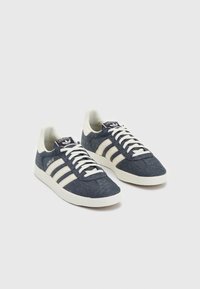 Blå textilsneakers med vita ränder, rundade tår, platta gummisulor och vita snören. Logotags på tungan. Slät textur.