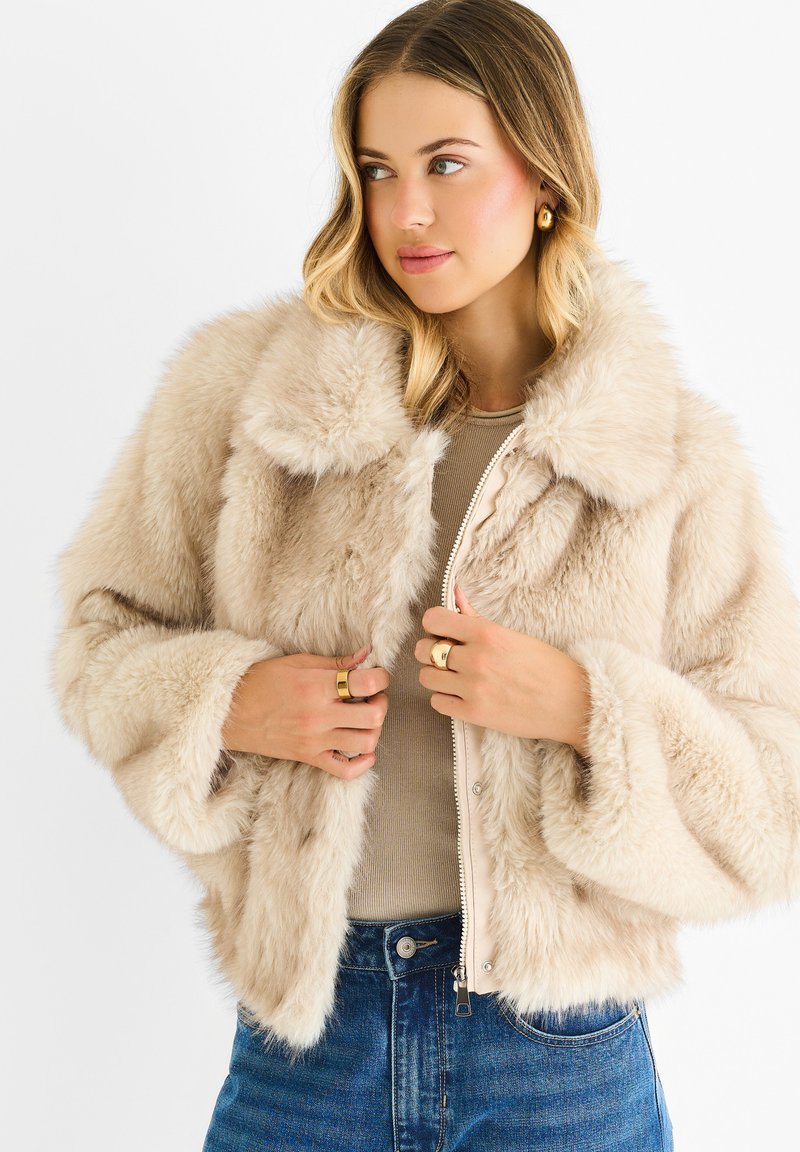 Woman with blonde hair wearing gold hoop earrings, gold rings, a beige faux fur jacket, beige top, and blue jeans against a white background.