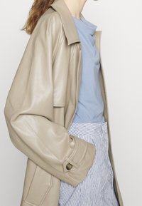 Beige läderjacka med avslappnad passform, med en vaksam krage och knappdetaljer. Bärs över en blå t-shirt och randiga byxor.