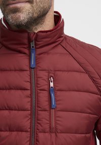 Giacca puffer borgogna con cuciture orizzontali, colletto alto e dettagli della cerniera blu. Presenta una piccola tasca con zip sul petto.