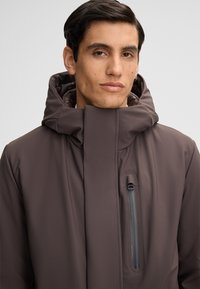 Bruine waterdichte jas met een hoge kraag, minimalistisch ontwerp en een ritsborstzak. Zachte voering zichtbaar rondom de kraag.