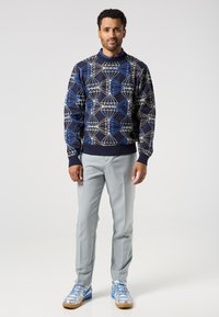 Man standing wearing a navy sweater with geometric blue and beige patterns, light gray trousers, and silver-blue sneakers.