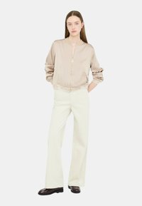 Giacca bomber in satin beige con zip frontale, vita elasticizzata con dettagli logo, indossata sopra pantaloni a zampa crema e scarpe scure.