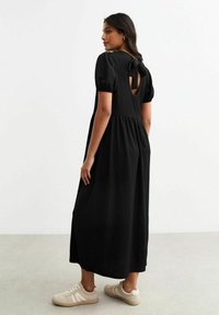 Abito midi nero con maniche a palloncino corte, dettaglio cutout sul retro e tessuto strutturato. Indossato con sneakers beige, in piedi su una superficie chiara.