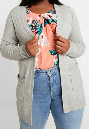 Vrouw draagt lichtgrijze cardigan over koraalkleurige top met bloemenprint en blauwe spijkerbroek, houdt met beide handen de randen van de cardigan dichtbij haar borst vast.