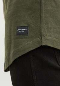 Olijfgroene katoenen T-shirt met een rechte zoom, voorzien van een zwart label met witte tekst "JACK & JONES" en "EST. 1990." Zwarte broek zichtbaar daaronder.