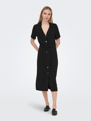 Femme debout portant une robe midi noire à boutons sur le devant, manches courtes et ceinture nouée à la taille, avec des chaussures noires faciles à enfiler.