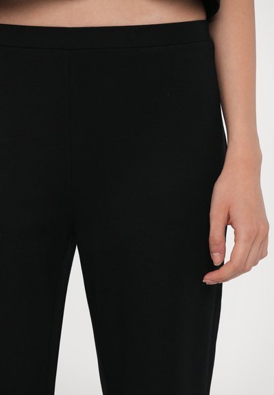 Schwarze dehnbare Hose mit glatter Textur, breitem Bund und schmalem Schnitt. Eine Hand ruht sanft an der Seite der Hose.