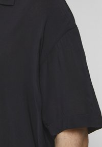 Camisa oscura y ligera con cuello estructurado y mangas cortas. Presenta una textura suave y detalles de costura sutiles.
