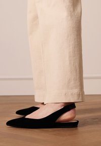 Zwarte fluwelen slingback balletjes met een spitse neus, gedragen met beige wijde broekspijpen, die een gladde textuur en minimalistisch ontwerp tonen.